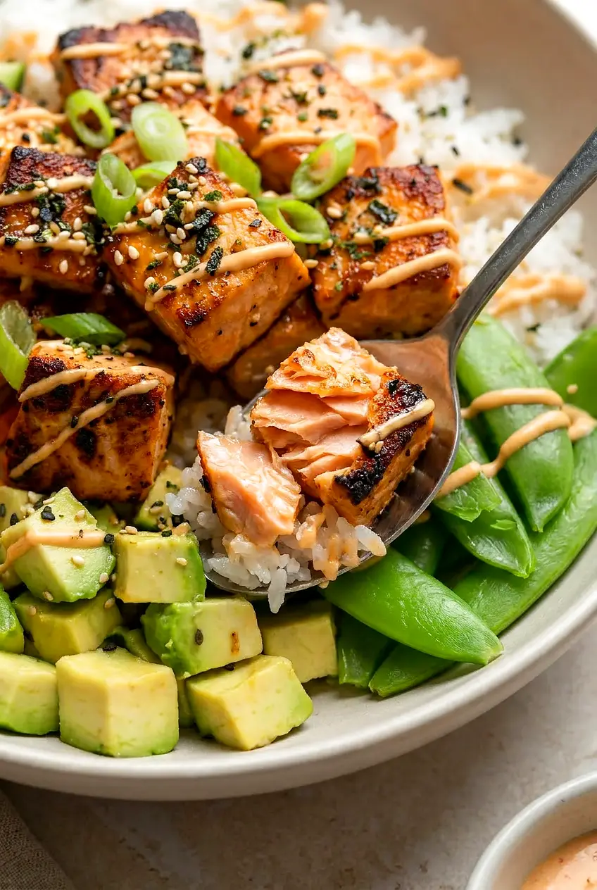 Close-up of flaky air fryer salmon cubes in a bowl with sriracha mayo drizzle, scallions, and sesame over rice.
