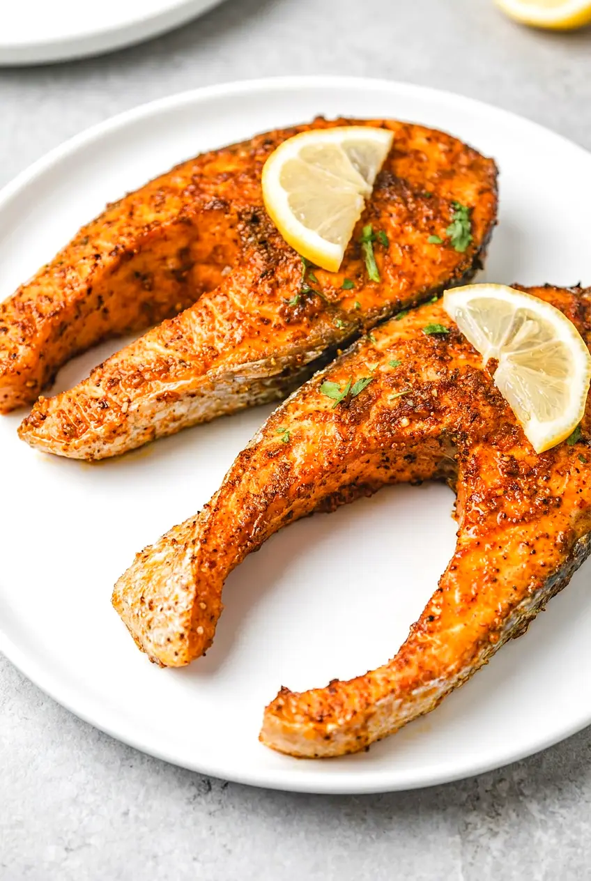 Air fryer salmon steak with Old Bay seasoning, lemon wedges, and light parsley on a white plate, browned edges and flaky texture