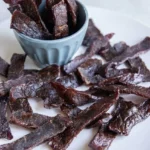 Air fryer beef jerky with dark, wrinkled marinated beef strips on a white plate and in a small blue bowl.